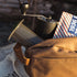 A backpack sitting outdoors with an Aeropress Brewer, Grinder and bag of coffee sticking out of the backpack.