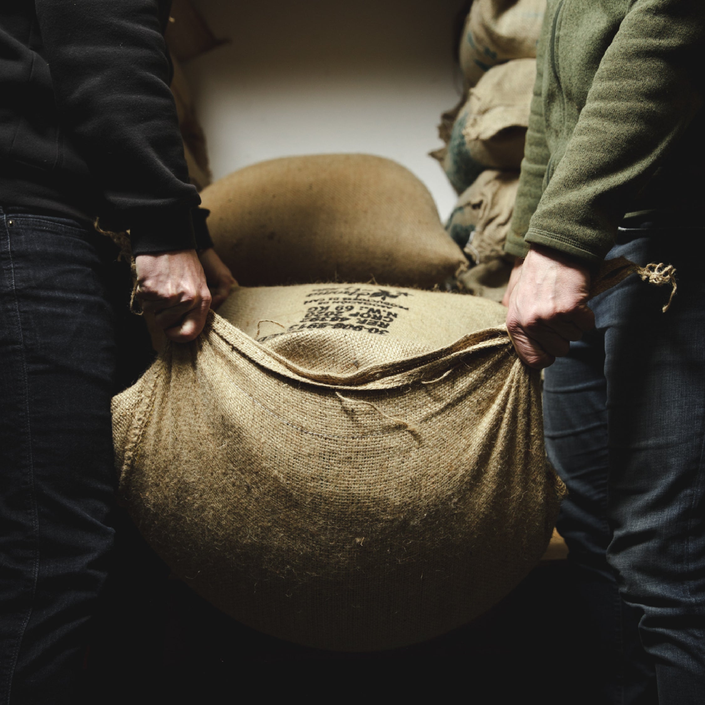Two people holding either side of a full burlap coffee sack in a roastery setting.