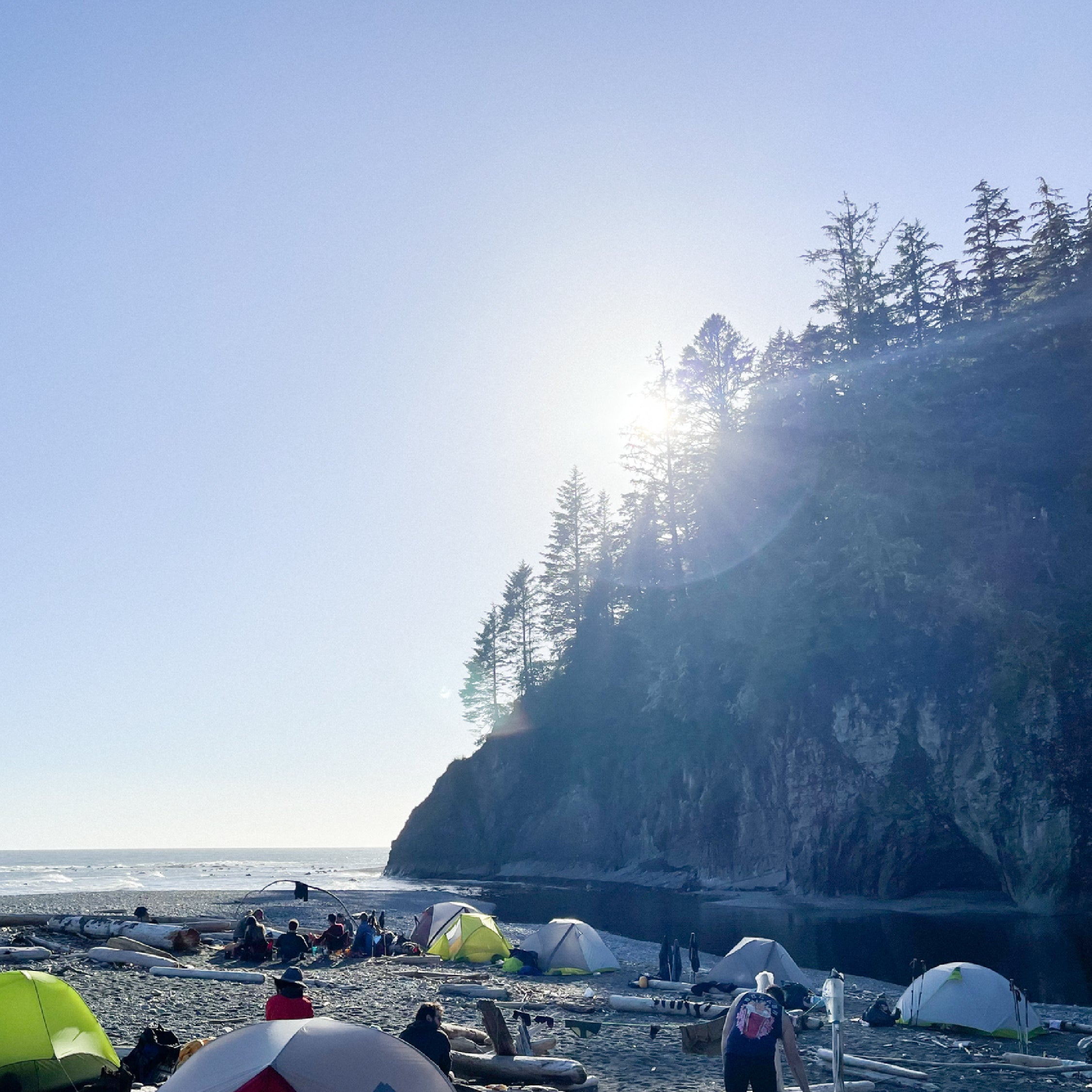 Tents and people on a beach on the West Coast Trail, sun breaking over a cliff.