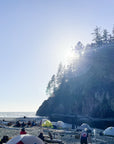 Tents and people on a beach on the West Coast Trail, sun breaking over a cliff.